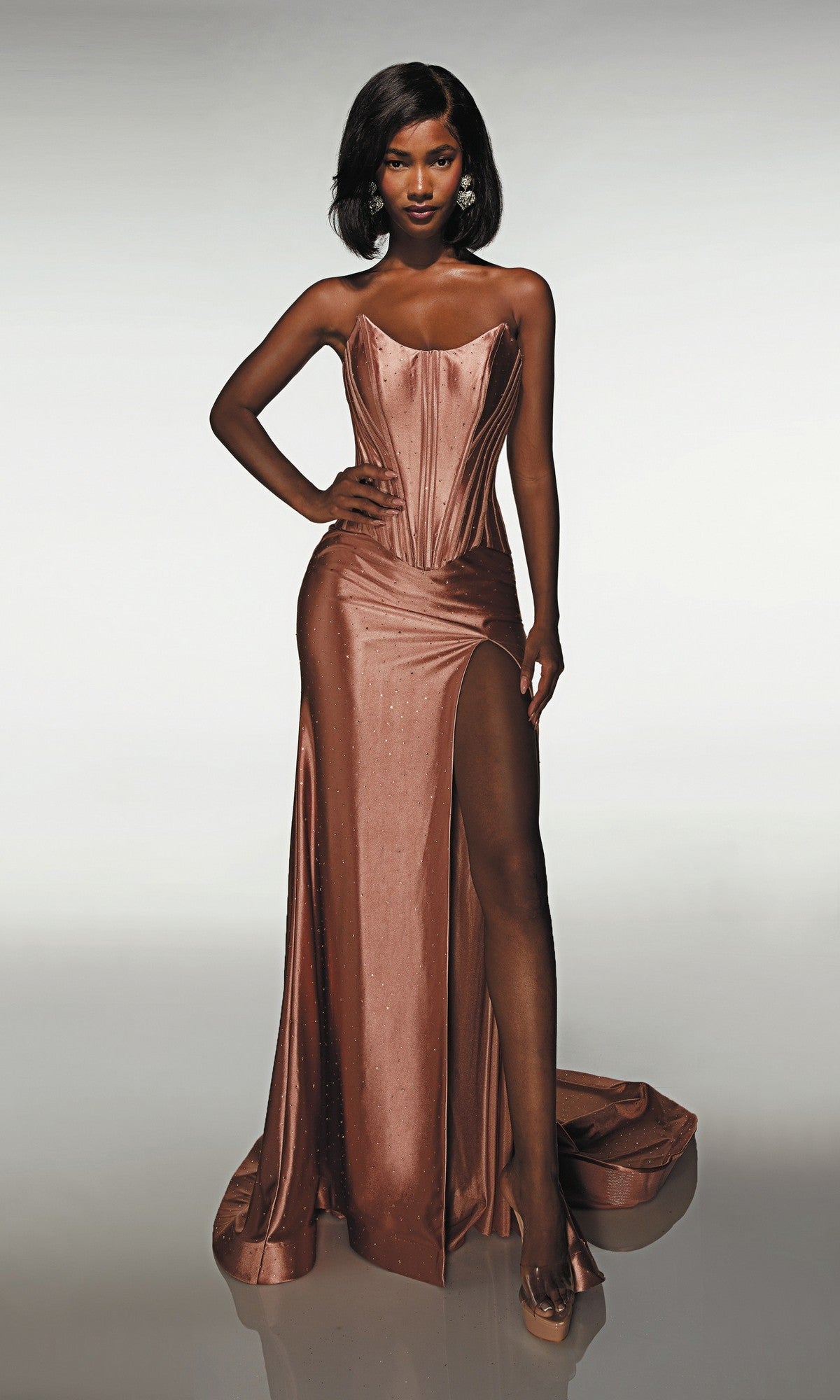 A woman poses with one hand on her hip in front of a plain backdrop, wearing the Alyce 62094 long prom dress—a strapless, form-fitting satin copper gown with a corset bodice and high thigh slit.